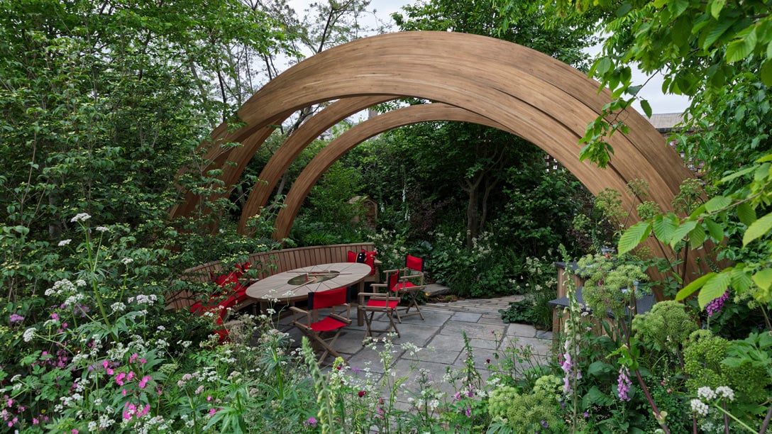 Chelsea Pensioner Archway featuring lush green plants and trees with a central wooden archway rising above a wooden bench and table with iconic scarlet cushions