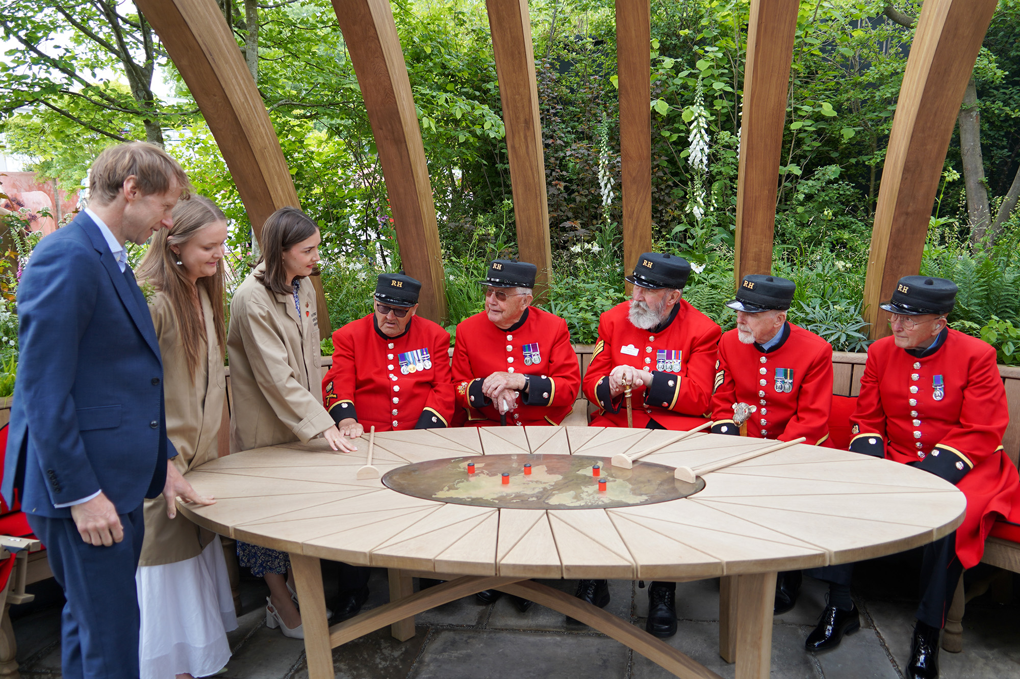 Chelsea Pensioners sit on a bench in front of a round wooden table with a map and markers showing places they have served. Above is a wooden archway and behind are lush green plants and trees.