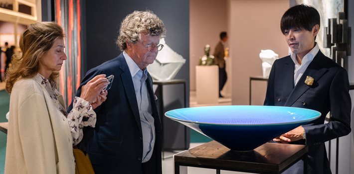 Two men and a lady stand behind a giant coral blue vase balanced upon a wooden plinth