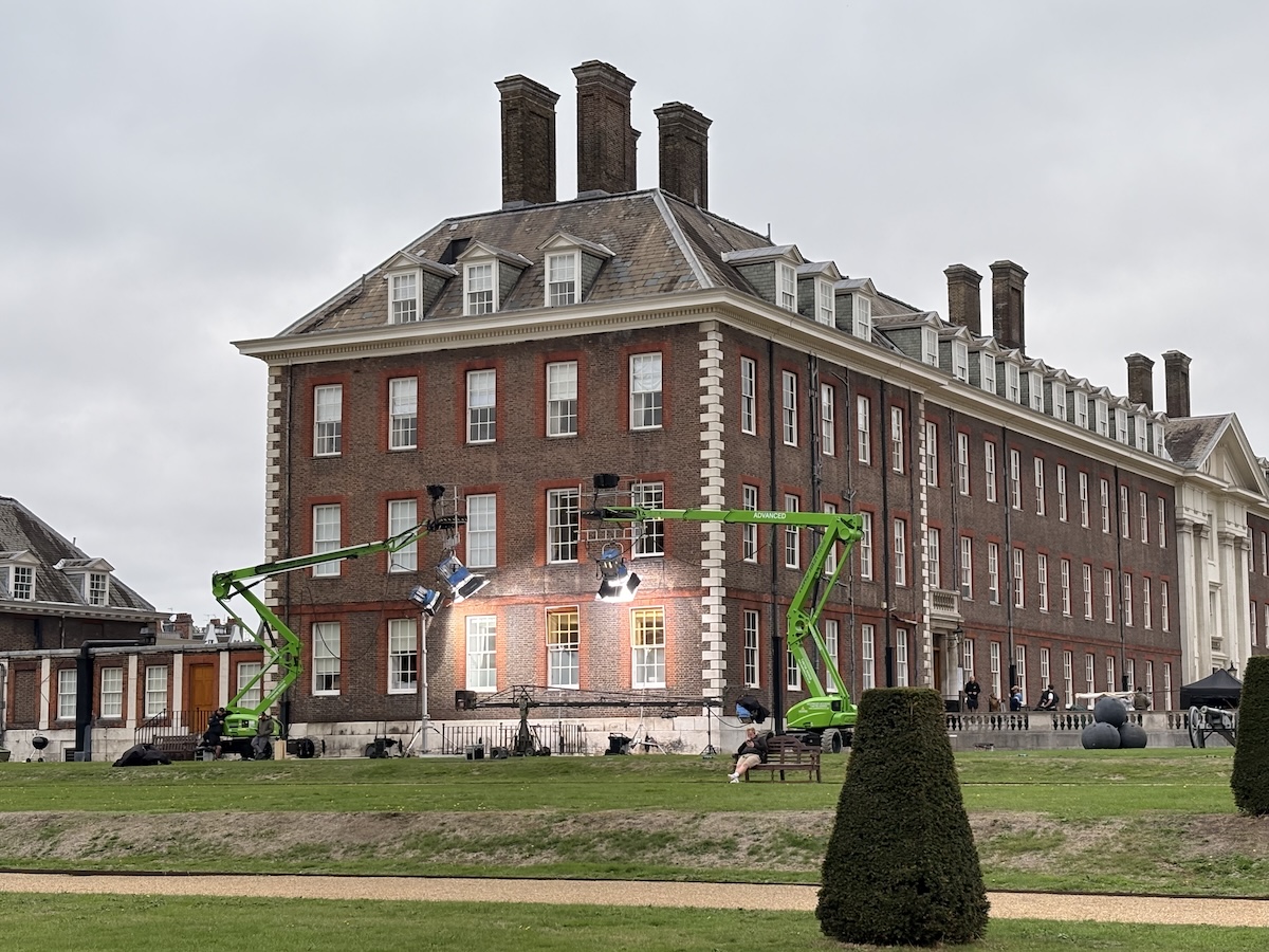 A film crew is working outside a large red brick building at the Royal Hospital Chelsea. Two green mobile lifting platforms hold powerful studio lights aimed at the first floor windows, illuminating the facade. Crew members and equipment are positioned along the terrace, with a few people watching nearby. The foreground shows neatly kept lawns and shaped hedges under an overcast sky.