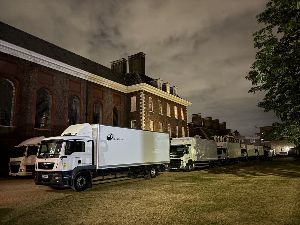 A row of large white production trucks is parked at night beside a red brick building at the Royal Hospital Chelsea. The building’s tall arched windows and white stone detailing are illuminated by exterior lighting. The sky is overcast and softly lit, and a tree frames the right side of the image. The trucks line the edge of a grassed area, suggesting an active film or television shoot taking place on site.