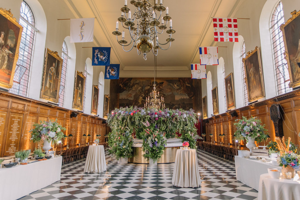A grand historic hall with black and white chequered flooring, tall arched windows, and wood panelled walls engraved with names. Large portraits in gilded frames line both sides of the room. Colourful heraldic banners hang from the ceiling. In the centre stands an elaborate floral installation with cascading greenery and vibrant flowers surrounding a candelabra. Several round cocktail tables draped in white cloth are arranged around the space, along with buffet tables decorated with floral arrangements. A l