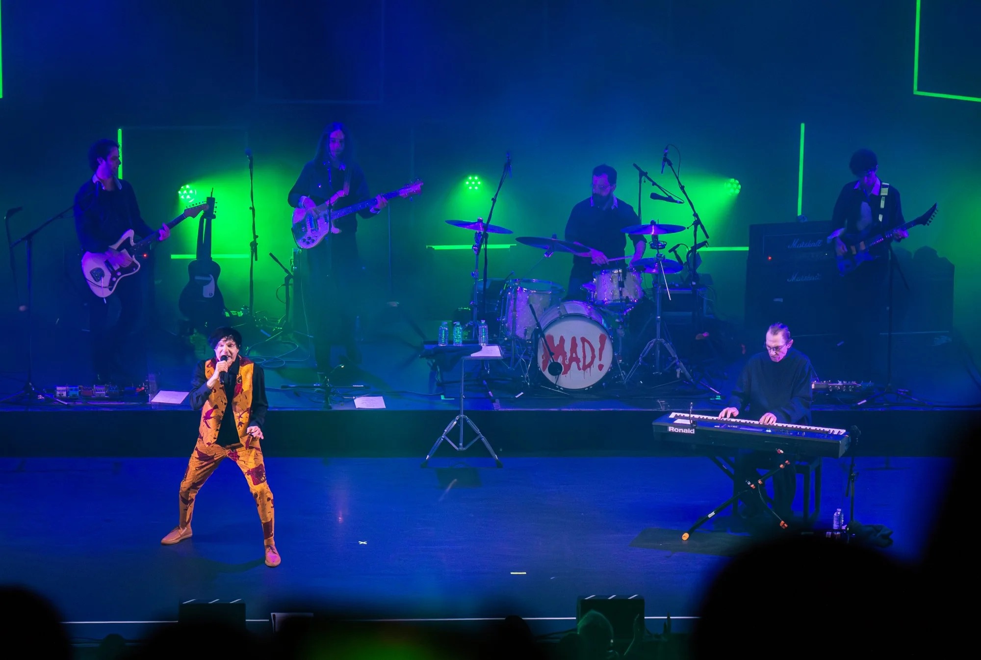 A band performs on a theatre stage under blue and green lighting. At the front, the lead singer stands mid-song wearing a bright patterned suit, while a keyboardist plays to the right. Behind them, guitarists and a drummer perform on a raised platform, with the drum kit marked “MAD!”. The scene conveys an energetic live concert atmosphere.