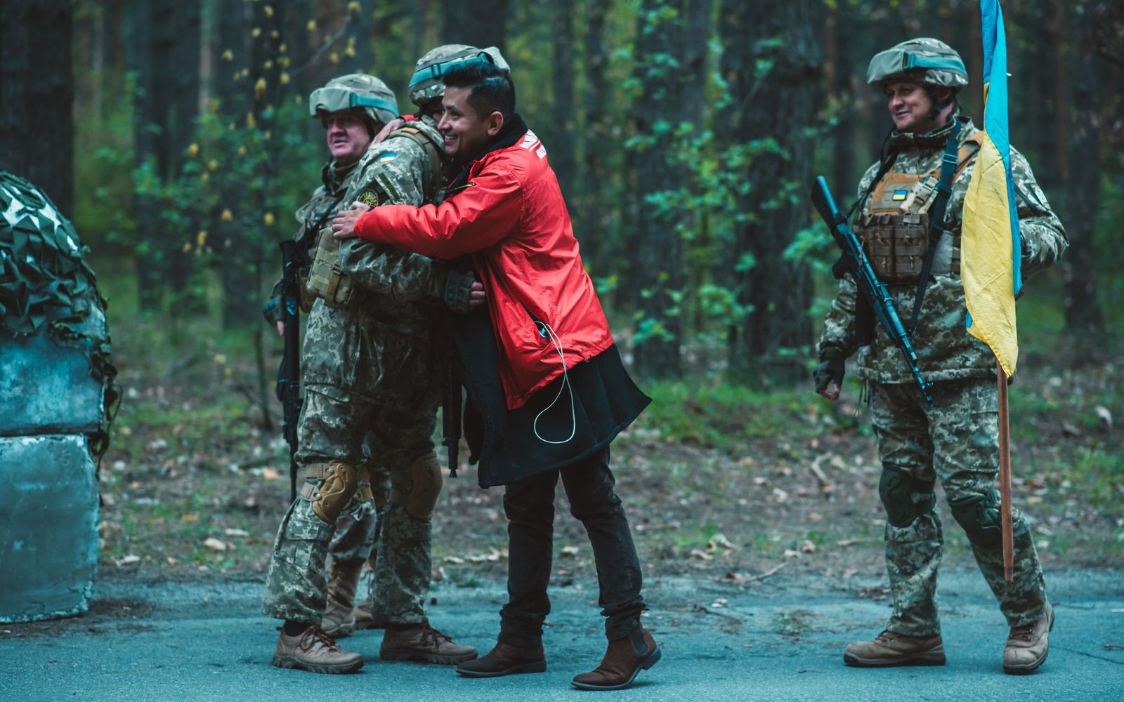 A civilian wearing a red jacket embraces a soldier in camouflage uniform on a forest road. Other armed soldiers stand nearby, one holding a blue and yellow flag. The scene conveys a moment of human connection and support amid a military environment.