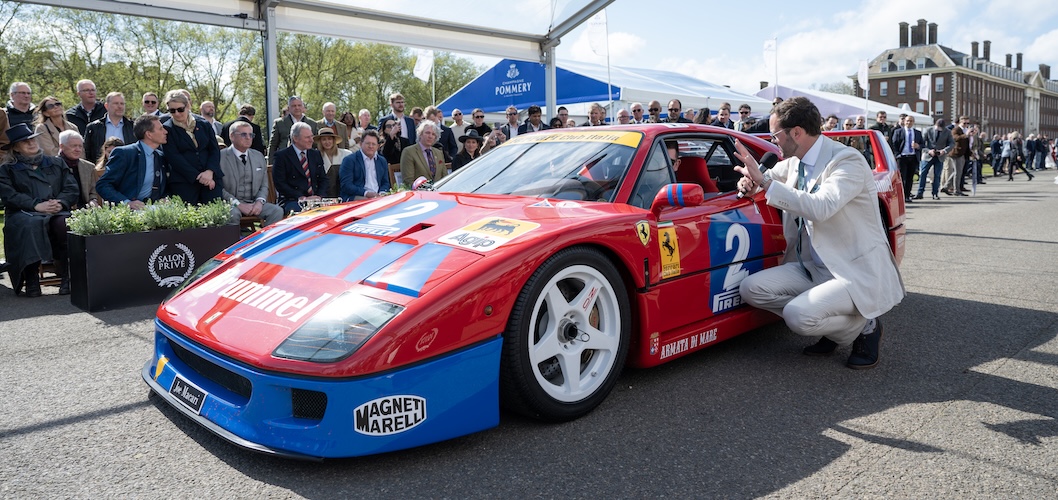 A driver in white overalls kneels down beside an open window of a red and blue sports car, with numerous adverts and logos positioned across the car. A large crowd of spectators is gathered behind the car.