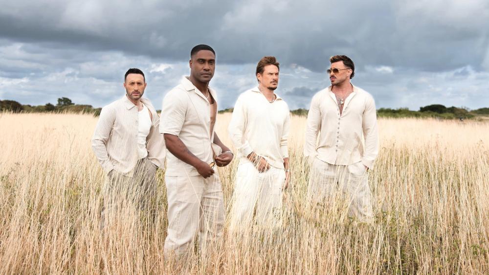 Four male performers stand in tall, golden grass under a dramatic cloudy sky, wearing coordinated light-coloured outfits. They face the camera with calm, confident expressions in an open countryside setting, creating a polished promotional-style image.