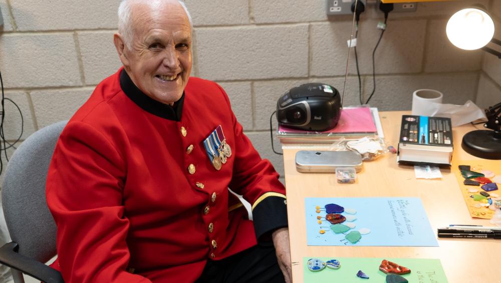 A Chelsea Pensioner wearing a scarlet uniform with service medals sits at a desk smiling towards the camera. In front of him are colourful pieces of glass and artwork arranged on paper, alongside craft tools and a desk lamp. The setting appears to be a workshop or studio space, suggesting a relaxed and creative activity.