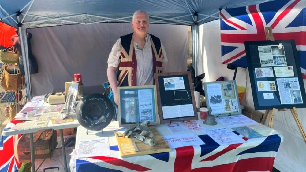 A man stands behind a display table under a canopy at an outdoor event. The table is covered with a Union Jack cloth and holds framed historical documents, photographs, artefacts, and information panels. A Union Jack flag hangs behind the display, suggesting a public history or object-handling exhibit.