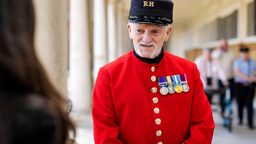 A Chelsea Pensioner in scarlet uniform with medals speaks to a visitor in a colonnaded walkway at the Royal Hospital Chelsea.