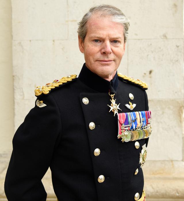 A highly decorated 4-star General of the British Army stands with hands behind his back against a white brick building