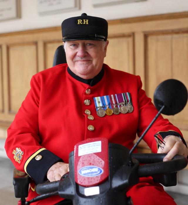 A Chelsea Pensioner in the traditional scarlet uniform and black RH shako hat sits on a mobility scooter, smiling warmly. Several service medals are pinned to his chest. The background shows wooden panelling and memorial plaques inside the Royal Hospital Chelsea.