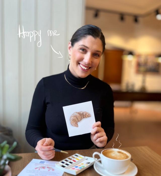 Woman smiling at a café table holding a small painting of a croissant, with watercolours and a cup of coffee in front of her.