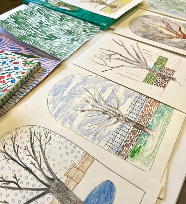 A selection of hand-drawn trees are displayed on a table underneath a colourful array of watercolour paintings.