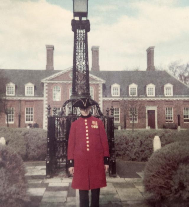 A vintage photo of a Chelsea Pensioner in iconic scarlet uniform with a black tricorn hat, standing within a paved garden with bushes all around him. A Christopher Wren building sits in the background, and rising up behind the Pensioner is an old black gated electric lamp post.