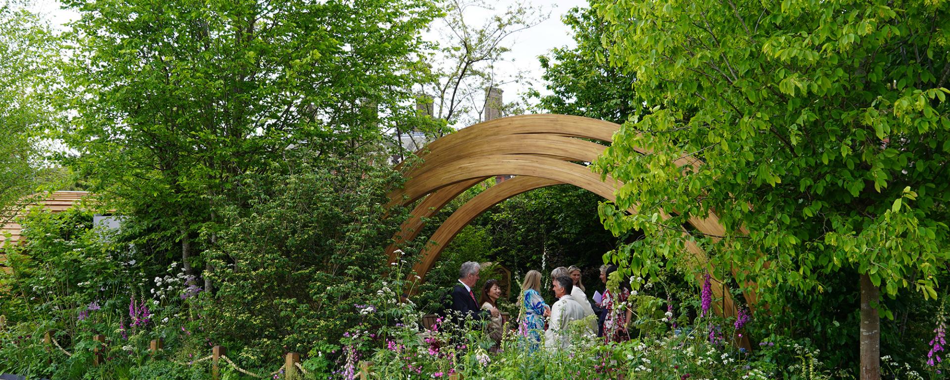 Gold award winning garden from London Square Developments and the Chelsea Pensioners - lush green leaves and trees surround a small group of people beneath a wooden archway,