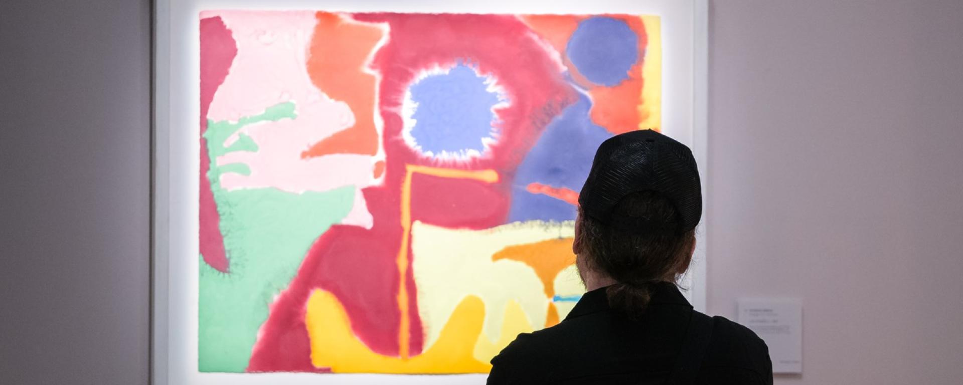 A silhouette of a man stands in front of a bold and vibrant art piece hanging on a contrasting grey wall
