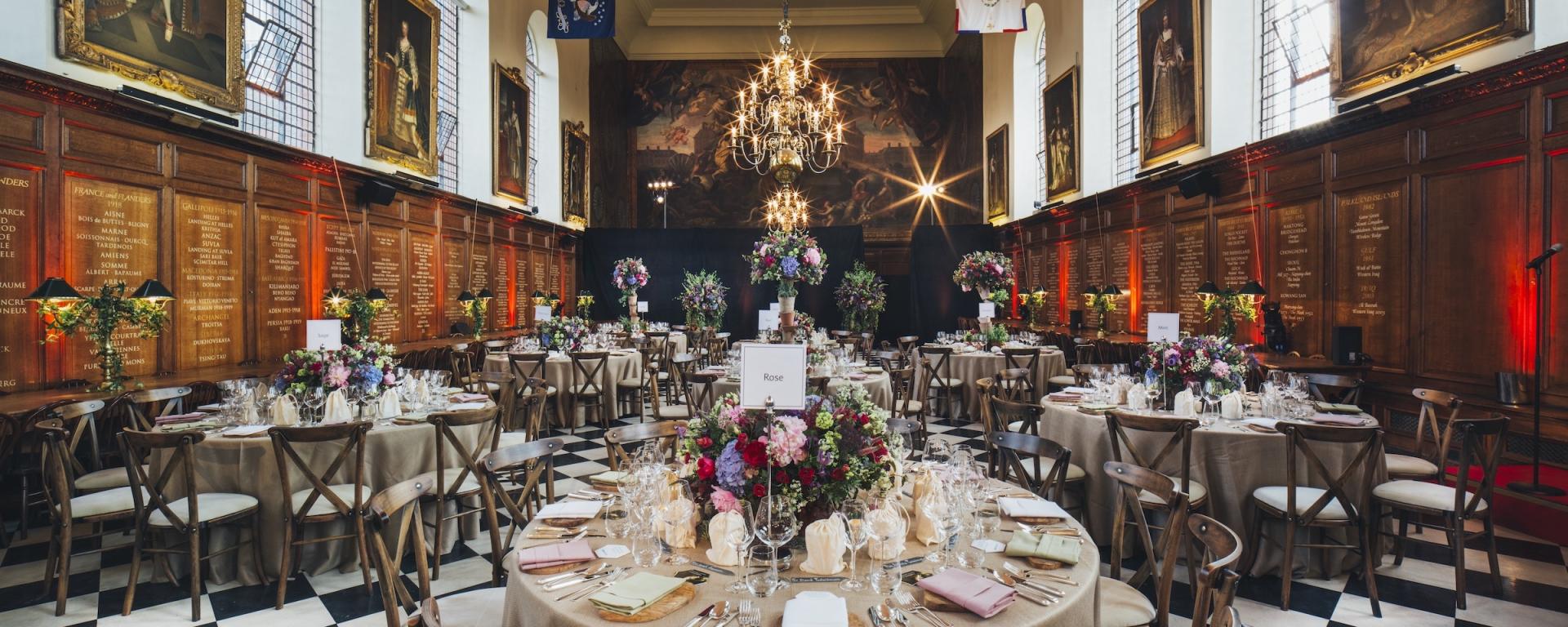 A grand historic hall arranged for a formal dinner with round tables covered in neutral linens, each decorated with large floral centrepieces. Chandeliers hang from the high ceiling, and portraits line the wooden panelled walls. The black and white chequered floor adds to the elegant setting.