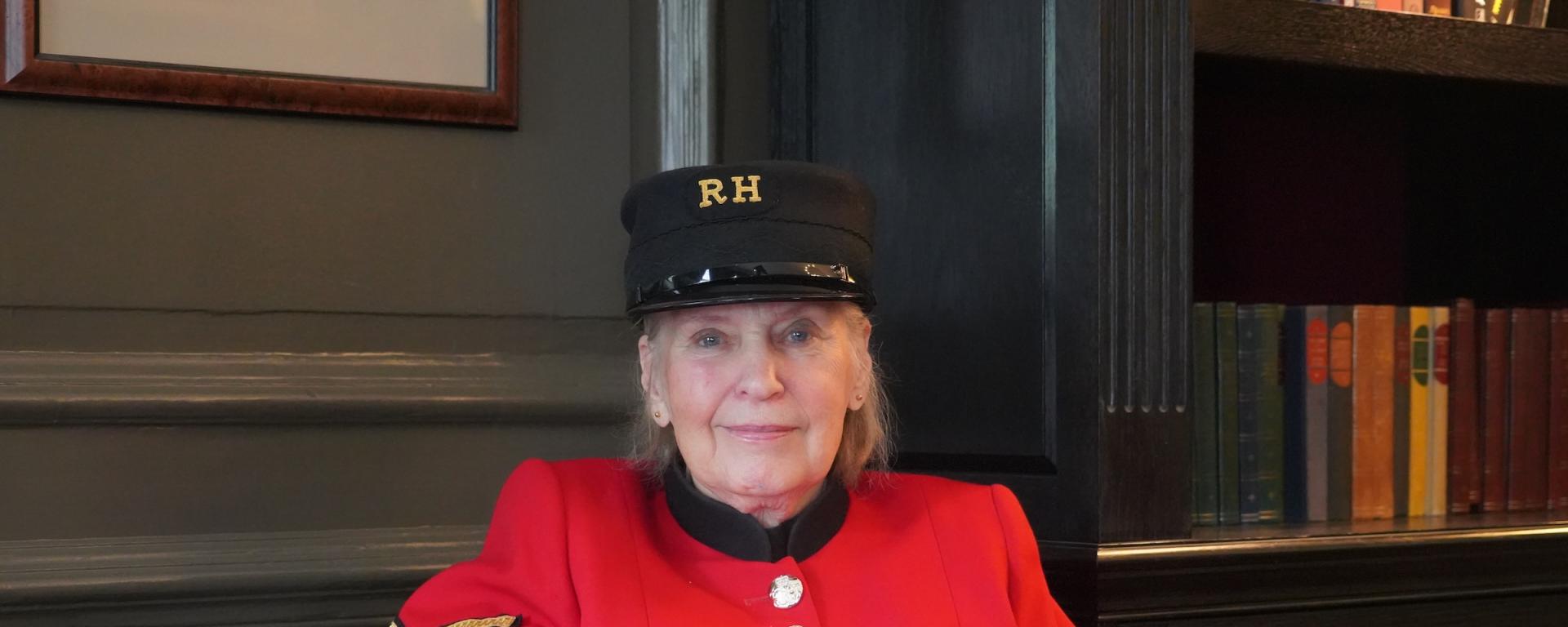 A lady Chelsea Pensioners sits within a wood panelled room in traditional scarlet uniform and black shako hat.