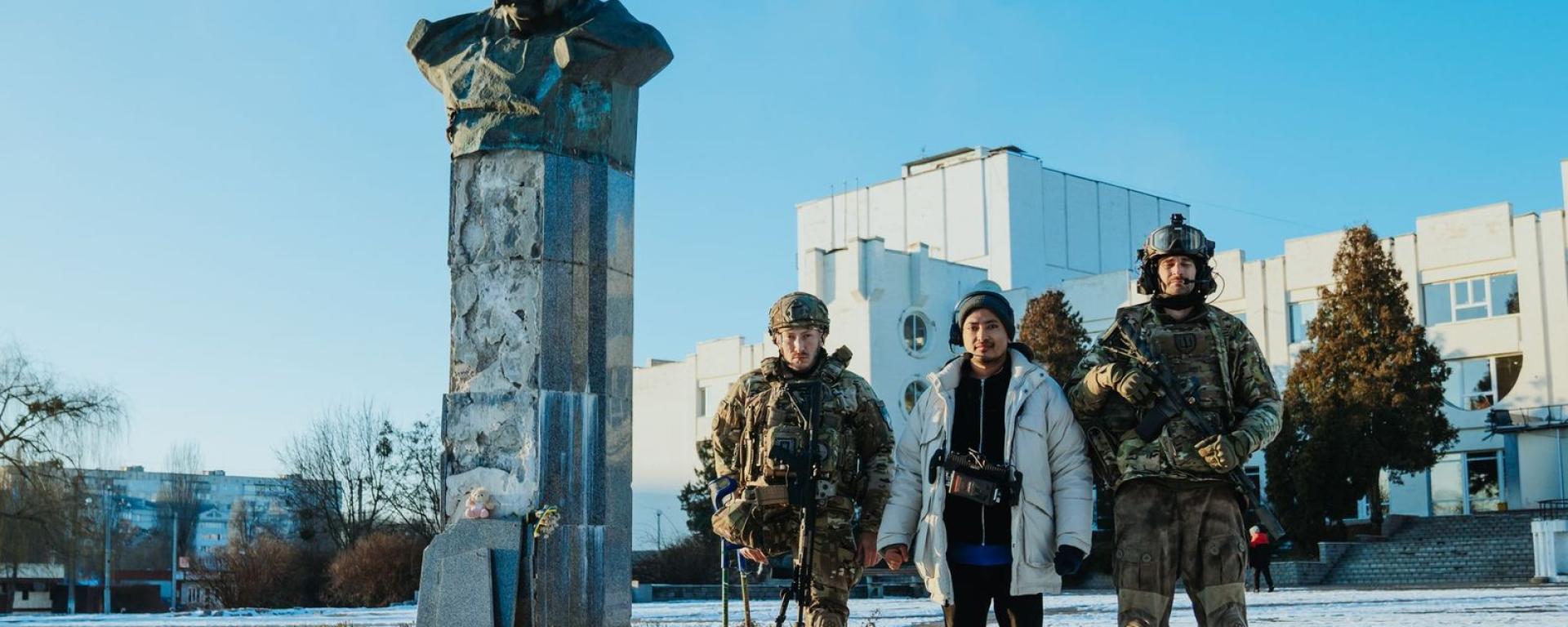 Three people stand in a snowy public square beside a weathered stone bust mounted on a pedestal. Two are soldiers in camouflage uniforms and helmets, while the person between them wears civilian winter clothing. A modern building stands in the background under a clear blue sky.