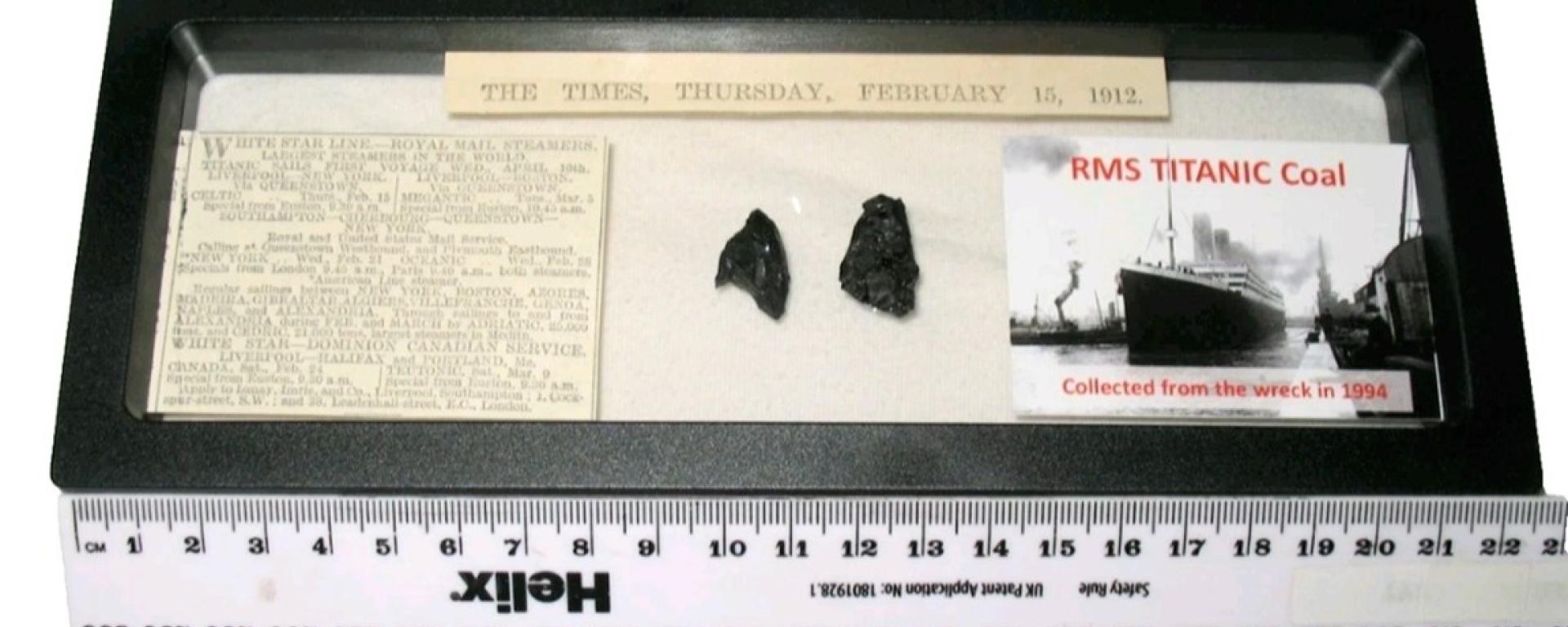 A framed display containing two small pieces of coal labelled “RMS Titanic Coal,” alongside a historic newspaper clipping dated February 15, 1912, and a photograph of the Titanic. A ruler beneath the frame shows the scale of the artefacts.