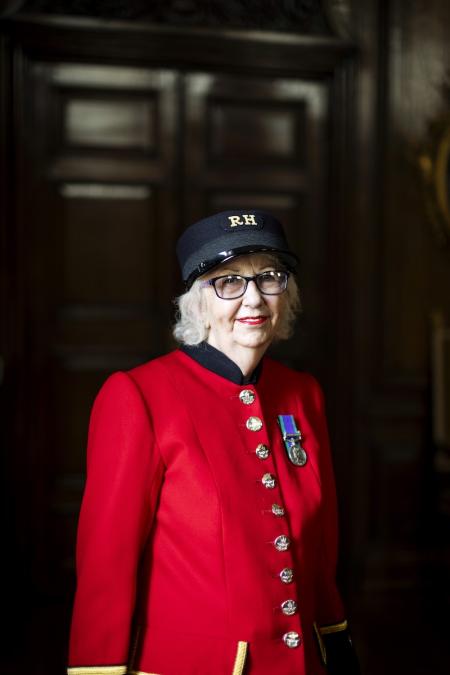 A female Chelsea Pensioner stands wearing her vibrant scarlet uniform, black shako hat and glasses, in front of large wood panelled doors.