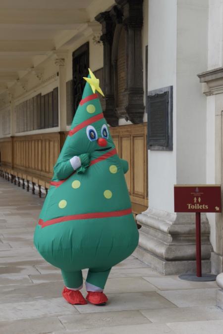 A person dressed in a large inflatable Christmas tree costume walks along an outdoor covered walkway at the Royal Hospital Chelsea. The green costume has a star on top, cartoon eyes, a red nose, and decorative spots and stripes. A sign pointing to the toilets stands nearby, and the background features historic stone and wood panelling.