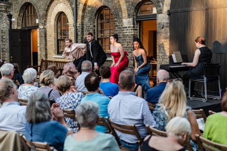 An outdoor opera performance takes place in a brick courtyard, with four singers in formal evening dress performing to a seated audience. A pianist plays at a keyboard to the side. Audience members sit closely together on wooden chairs, watching and applauding as the singers perform in front of arched doorways and windows.