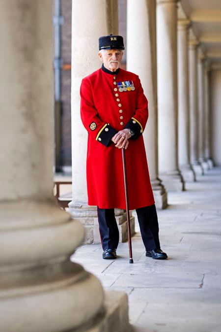 A Chelsea Pensioner in full scarlet uniform standing with a cane beneath the colonnade at the Royal Hospital Chelsea.