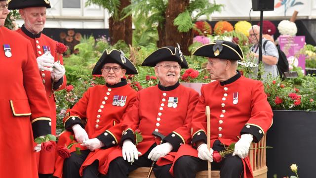 Three Chelsea Pensioners in Scarlet coats sat on a bench surrounded by flowers and plants