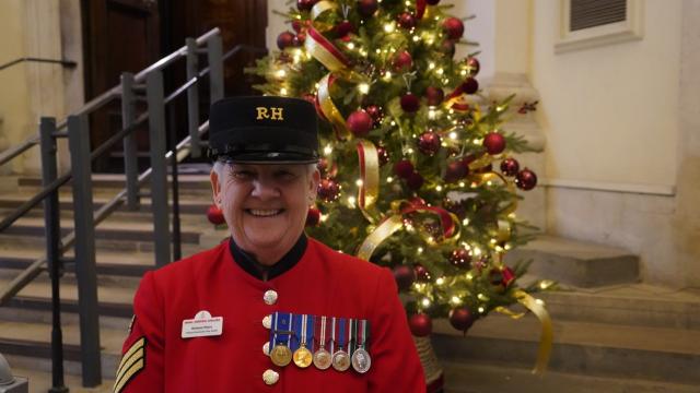A female Chelsea Pensioners stands in front of stone steps in the Royal Hospital Chelsea's Octagon foyer. Dressed in her famous Scarlet uniform and Shako hat, her name tag reads "Barbara - Tour Guide" her chest is adorned with half a dozen medals of various colours. Behind her a warmly decorated Christmas tree glitters in Red & Gold.
