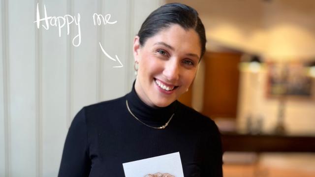 Woman smiling at a café table holding a small painting of a croissant, with watercolours and a cup of coffee in front of her.